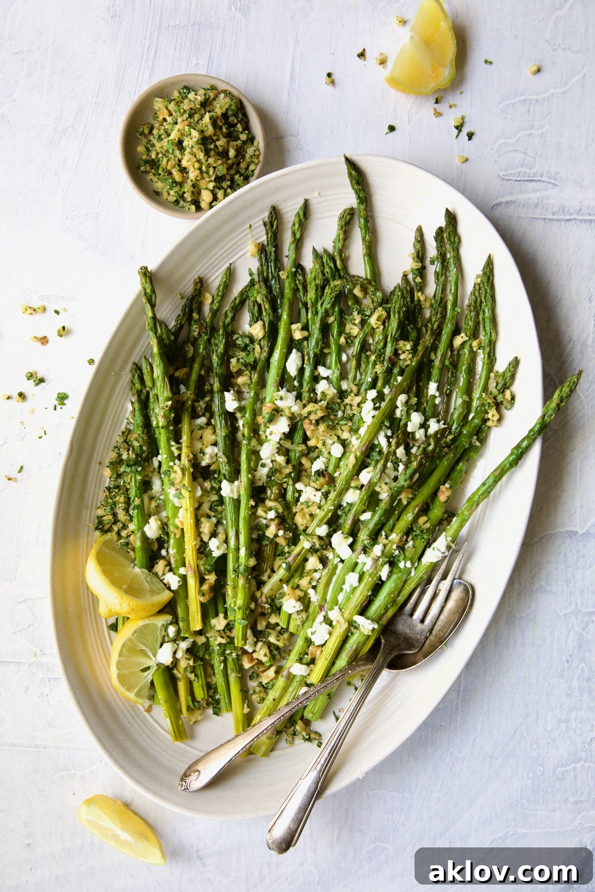 An enticing angled shot of Roasted Asparagus with Walnut Lemon Gremolata and Goat Cheese on a white oval platter, accompanied by lemon wedges, serving utensils, and a bowl of gremolata, highlighting its inviting presentation.