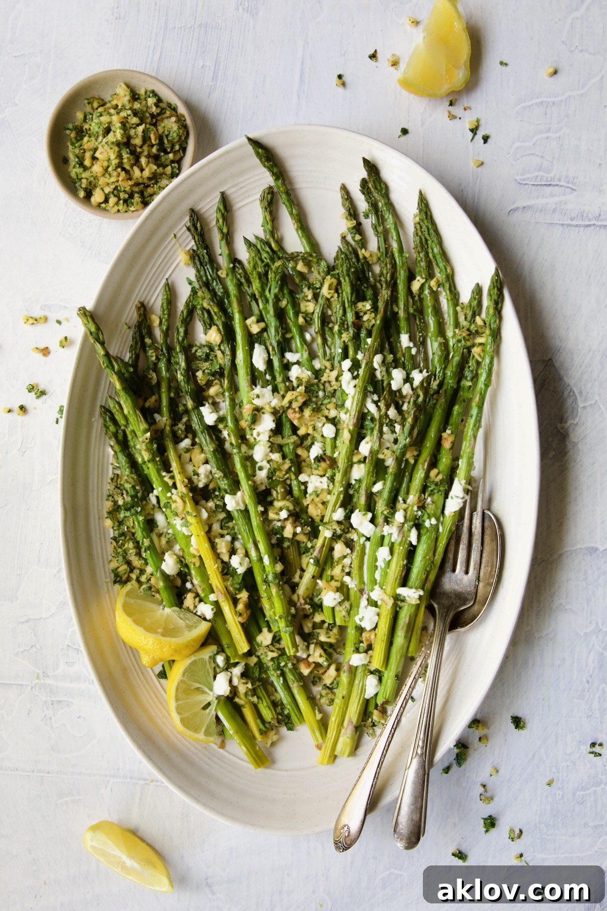 A vibrant serving of Roasted Asparagus with Walnut Lemon Gremolata and Goat Cheese, beautifully presented with lemon wedges, serving utensils, and a small bowl of gremolata for extra topping.