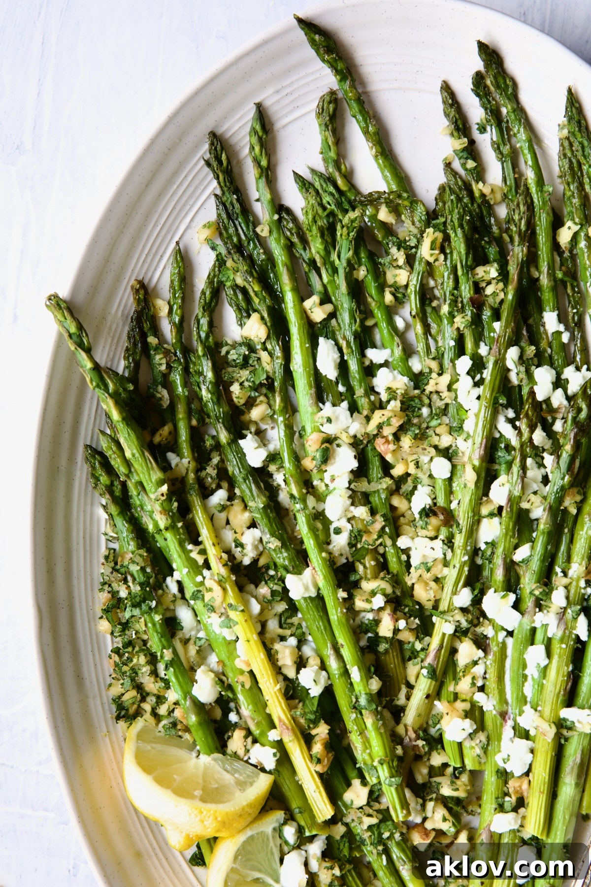 Roasted Asparagus with Walnut Lemon Gremolata and Goat Cheese on a white oval platter with fresh lemon wedges, ready to serve.