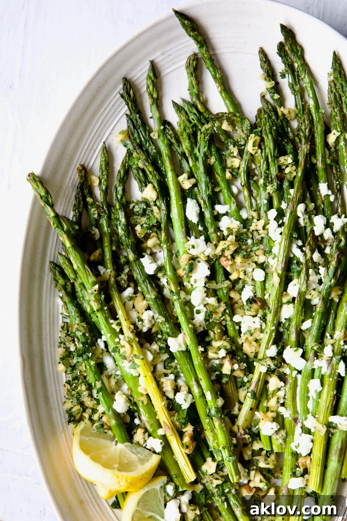 Close-up of Roasted Asparagus with Walnut Lemon Gremolata and Goat Cheese on an elegant white oval platter, with fresh lemon wedges beside it.