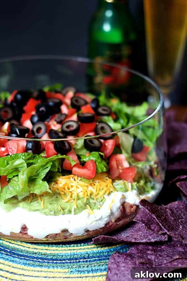 Vibrant Healthier 7-Layer Dip in a tall glass bowl, surrounded by blue corn chips and a refreshing beverage in the background, perfect for any gathering.