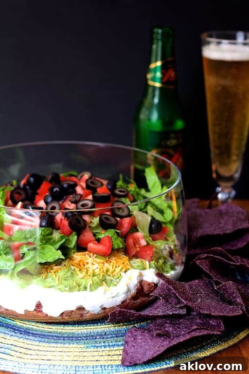 Close-up photo of the Healthier 7-Layer Dip in a glass serving bowl, placed on a colorful placemat, with scattered tortilla chips and a drink in the soft-focus background.