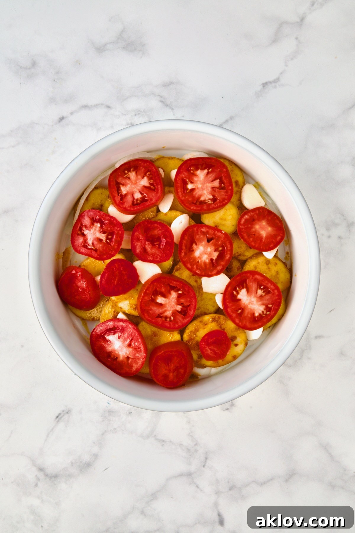 Tomatoes placed on top of potatoes, garlic and onions in round white baking dish.