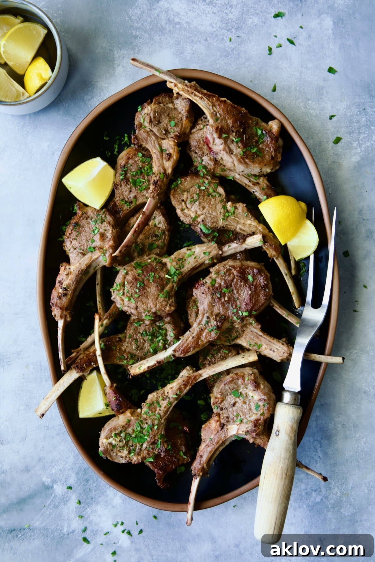 Greek Lamb Chops on black oval platter with meat fork garnished with lemon wedges.