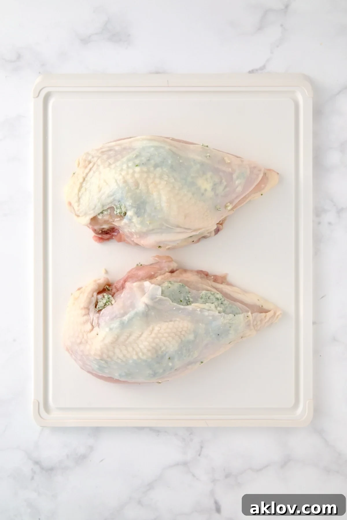 Two uncooked split chicken breasts on white plastic cutting board.