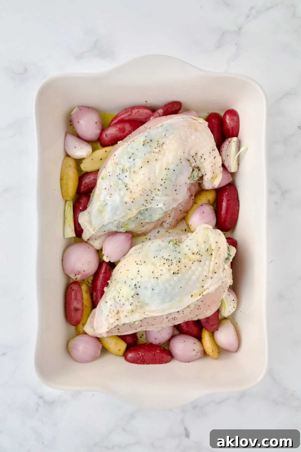 Uncooked potatoes and shallots in white rectangular baking dish with uncooked split chicken breasts placed on top.