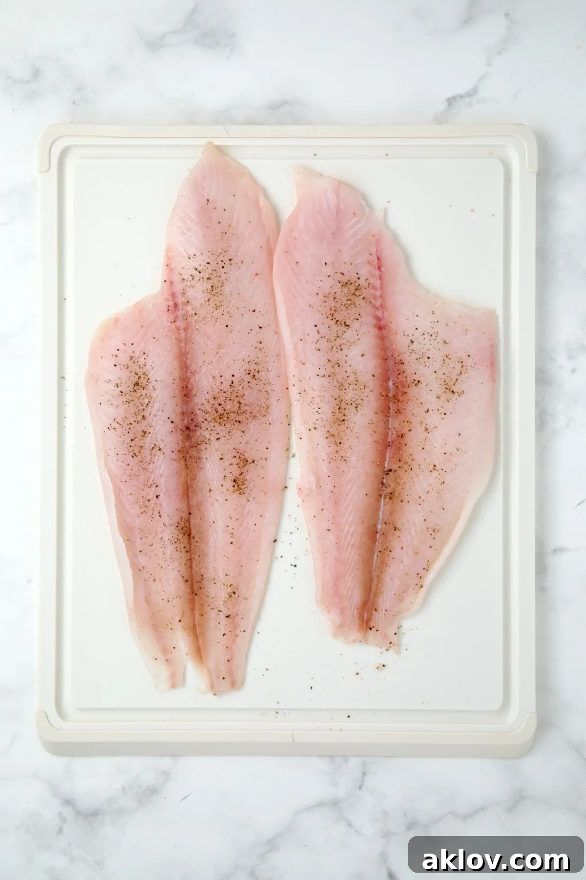 Two fresh, uncooked flounder fillets laid flat on a white plastic cutting board.