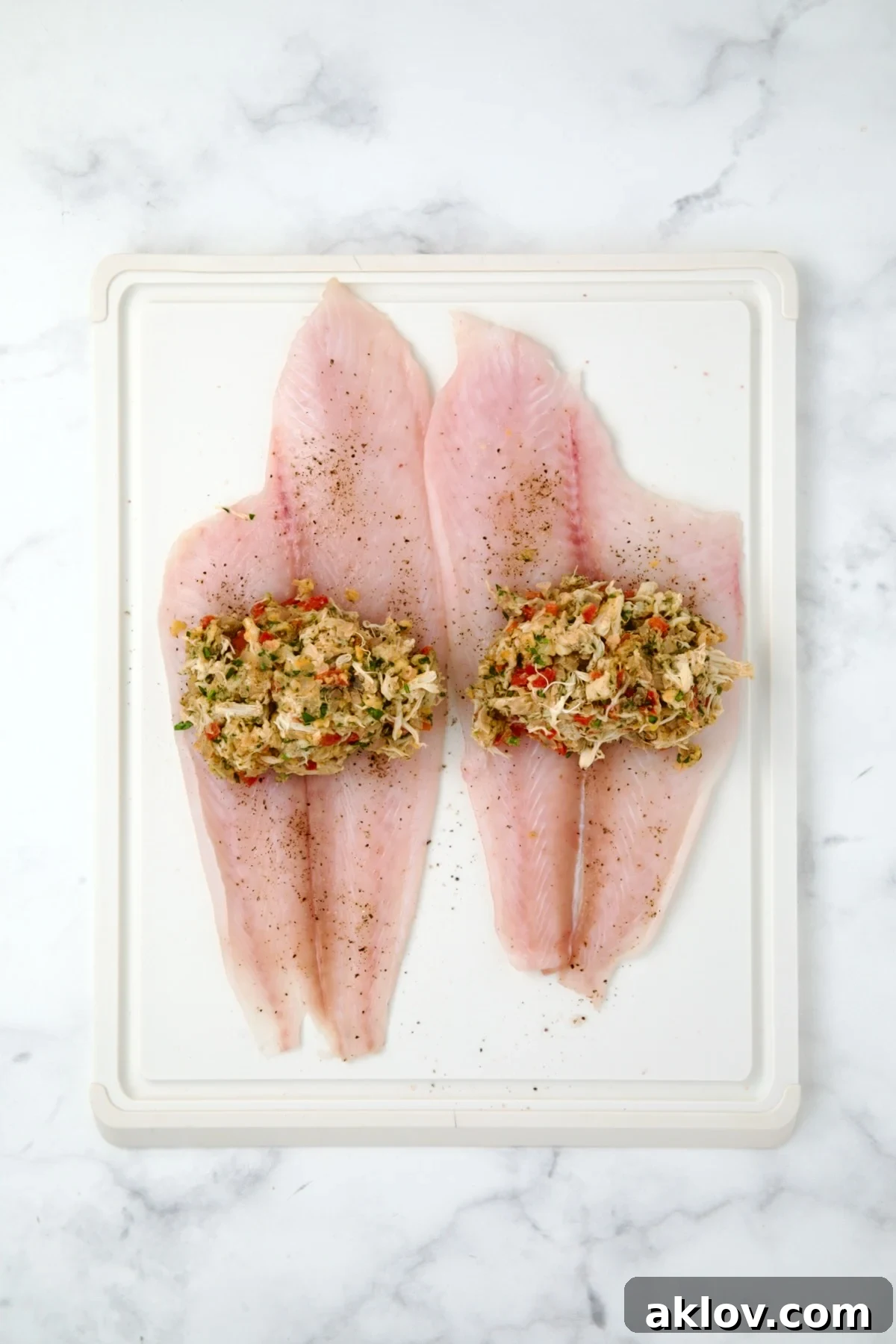Two uncooked flounder fillets on a white cutting board, each with a mound of crab filling placed in the center.