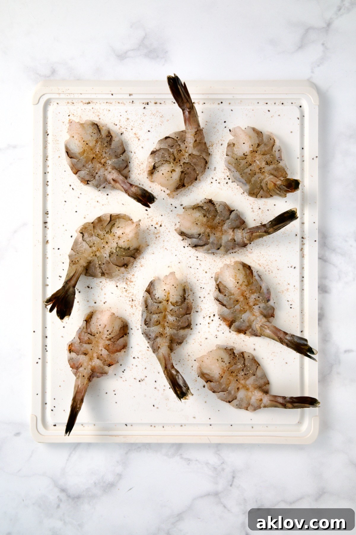 Nine colossal peeled and deveined shrimp on a white plastic cutting board, butterflied and seasoned with salt and pepper.