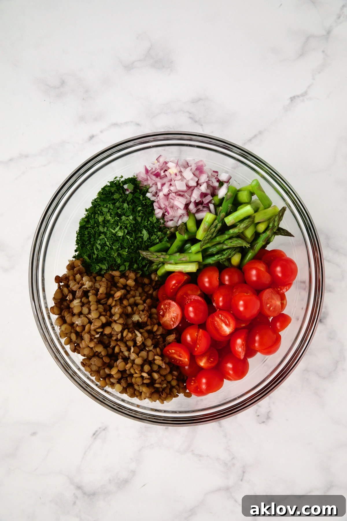 The base ingredients for Lemony Lentil Salad - cooked lentils, blanched asparagus, halved cherry tomatoes, finely chopped red onion, and fresh parsley - gathered in a large glass mixing bowl, prior to dressing and feta.