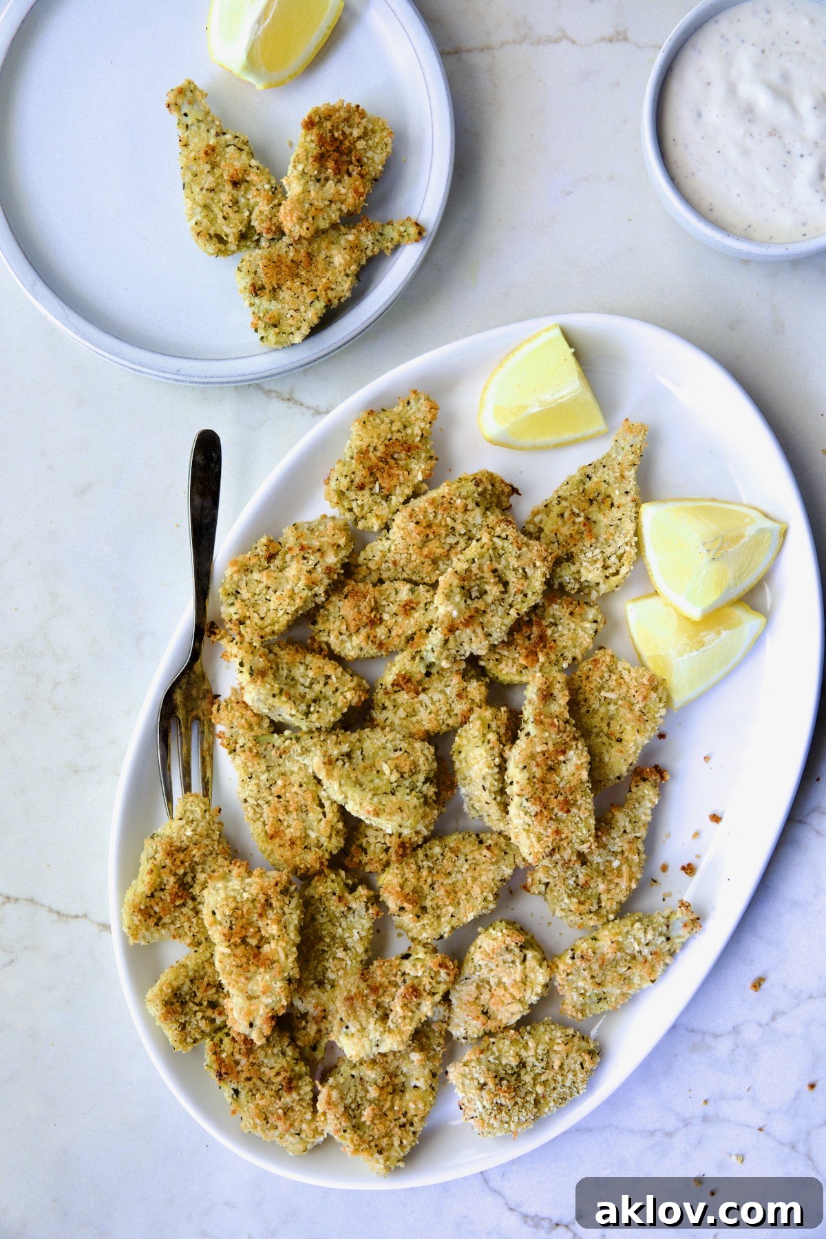 Crispy Parmesan Artichoke Hearts on oval white platter wiht some placed on a small plate with lemon wedges.