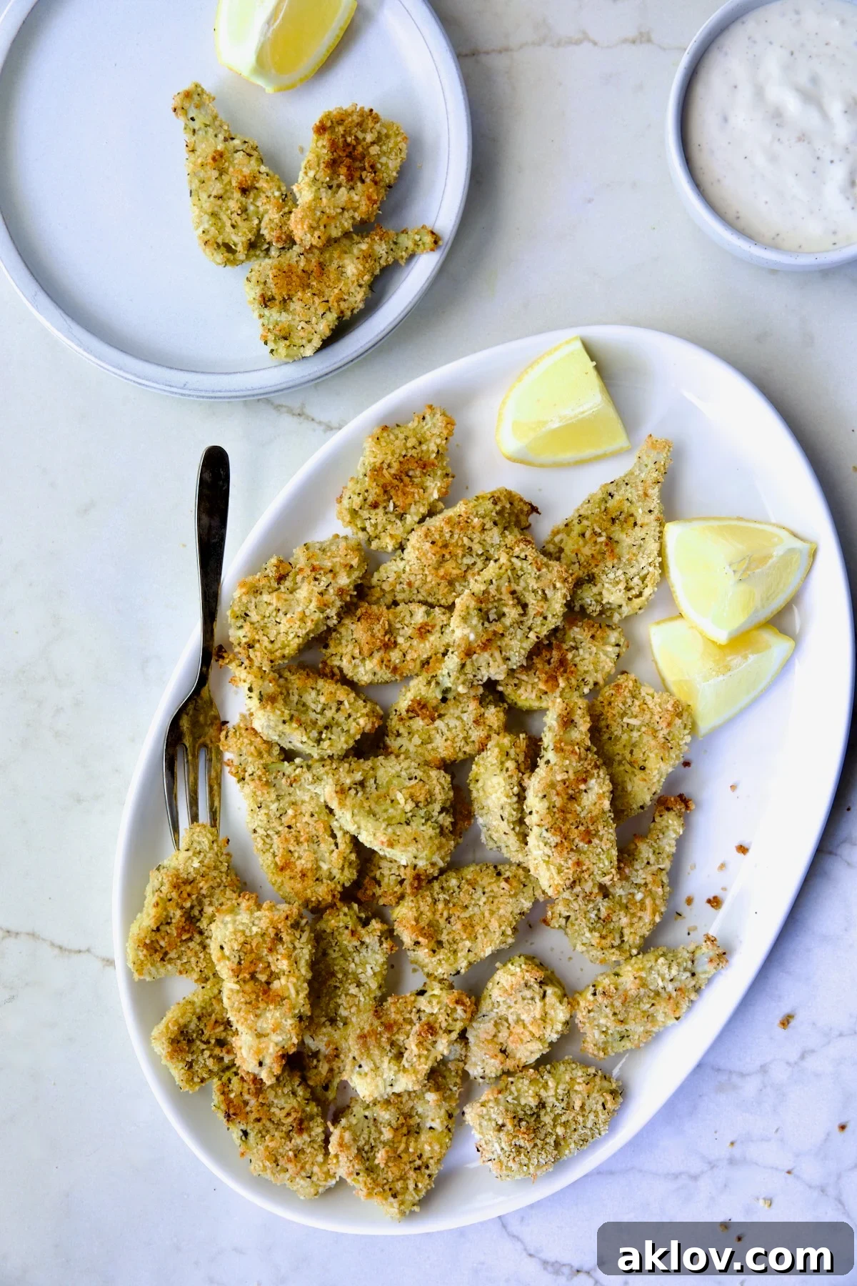 Crispy Parmesan Artichoke Hearts on oval white platter wiht some placed on a small plate with lemon wedges.