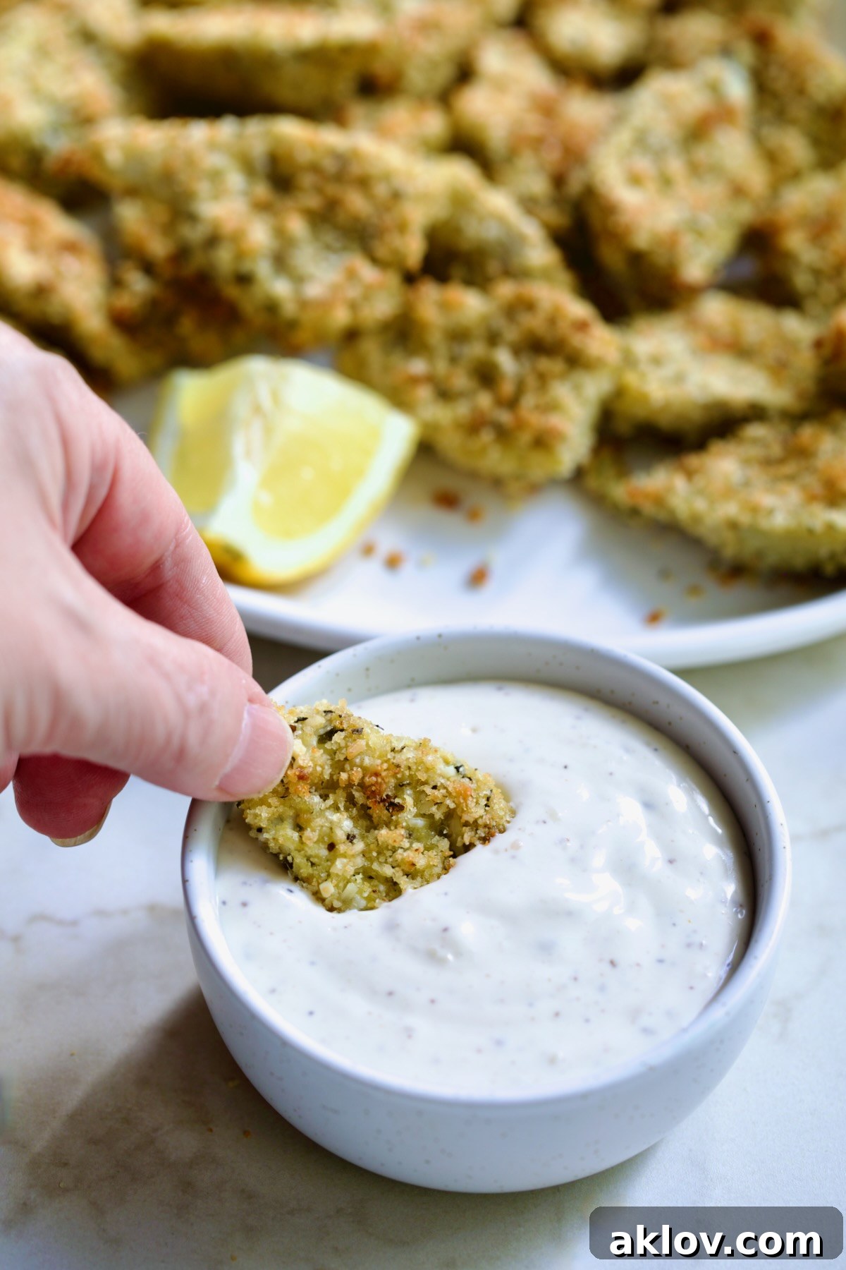 Hand dipping an artichoke heart into the Caesar dip in a white bowl.