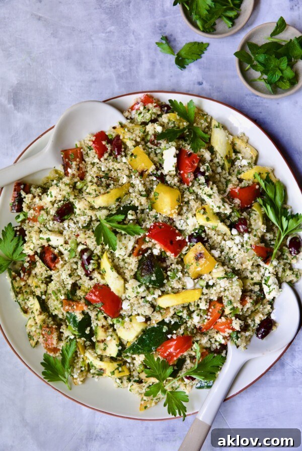 Mediterranean Quinoa Salad with Grilled Vegetables on round white platter with serving utensils garnished with herb sprigs.