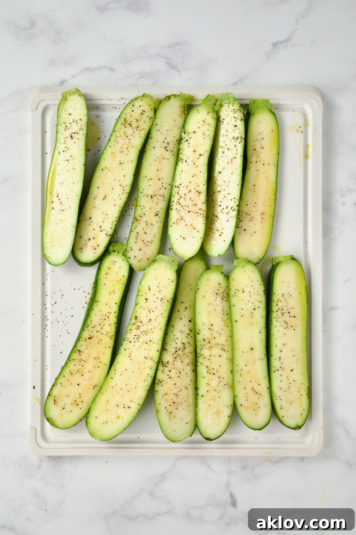 Sliced zucchini on white plastic cutting board seasoned with salt, pepper and drizzled with olive oil.