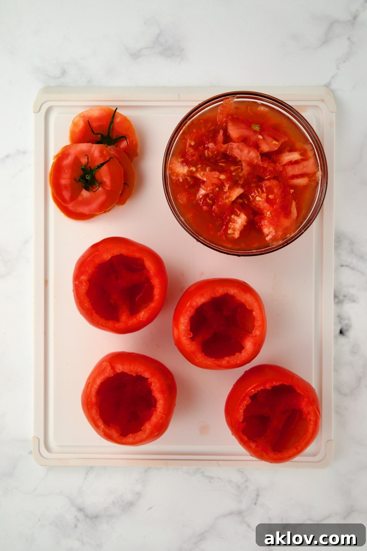 Four hollowed-out round tomatoes with the lids and the scooped-out pulp in a glass bowl on a white plastic cutting board.