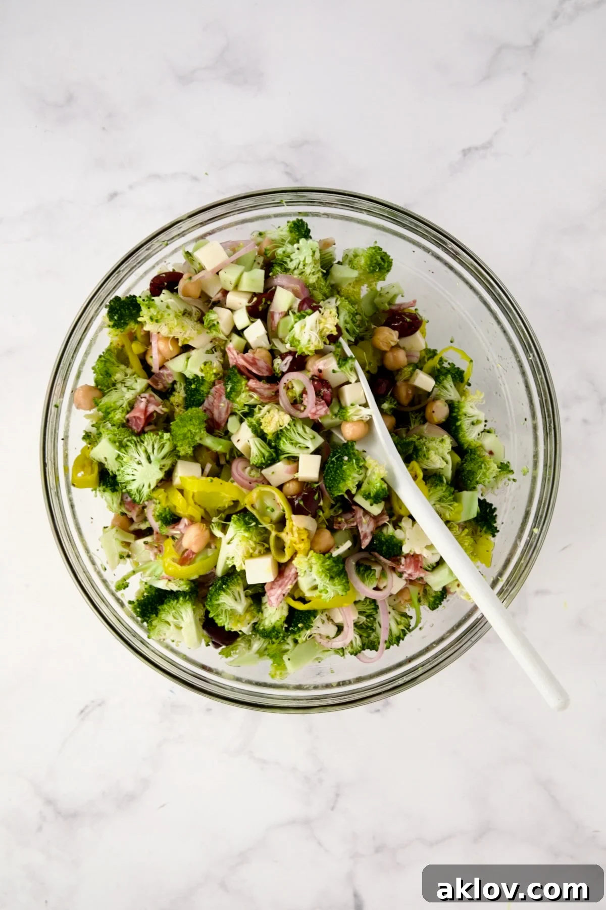 Ingredients for Italian Broccoli Salad being mixed together in glass bowl with white mixing spoon.