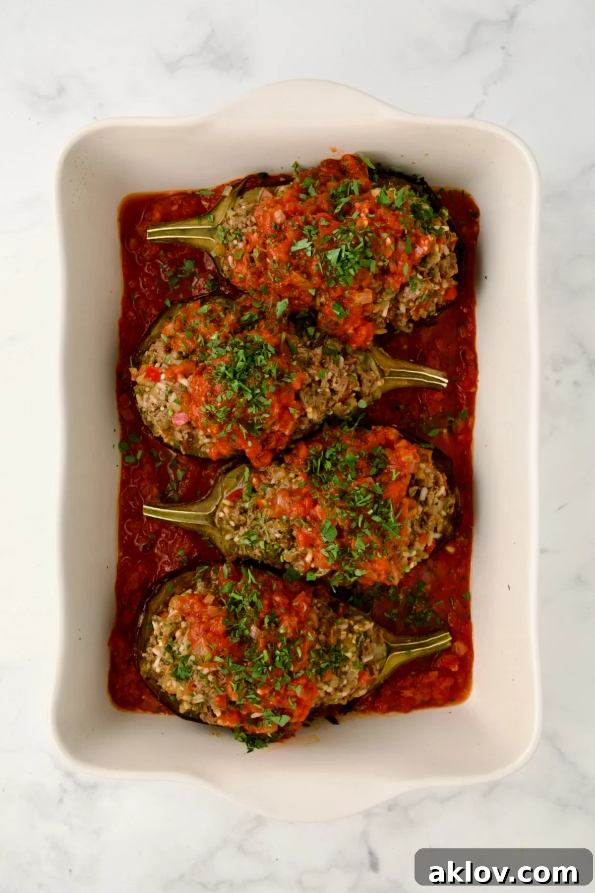 Four Stuffed Eggplant halves set a top tomato sauce in white rectangular baking dish topped with tomato sauce ready to be baked.