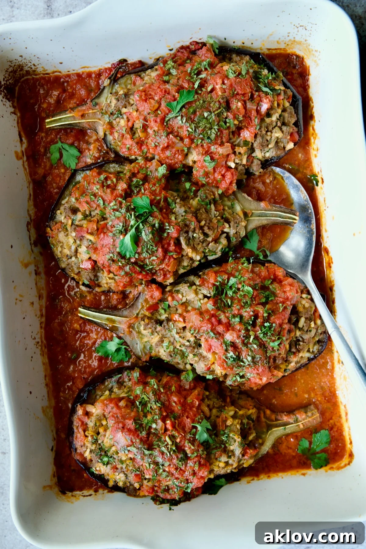 Four baked Stuffed Eggplant halves set a top tomato sauce in white rectangular baking dish with serving spoon.