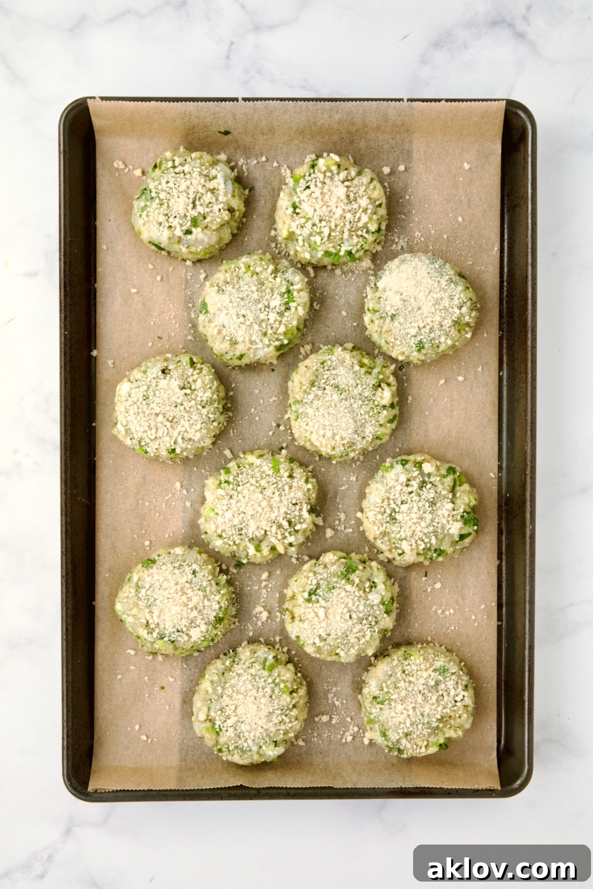 12 uncooked shrimp cakes on parchment paper lined rimmed baking sheet with panko sprinkled over.