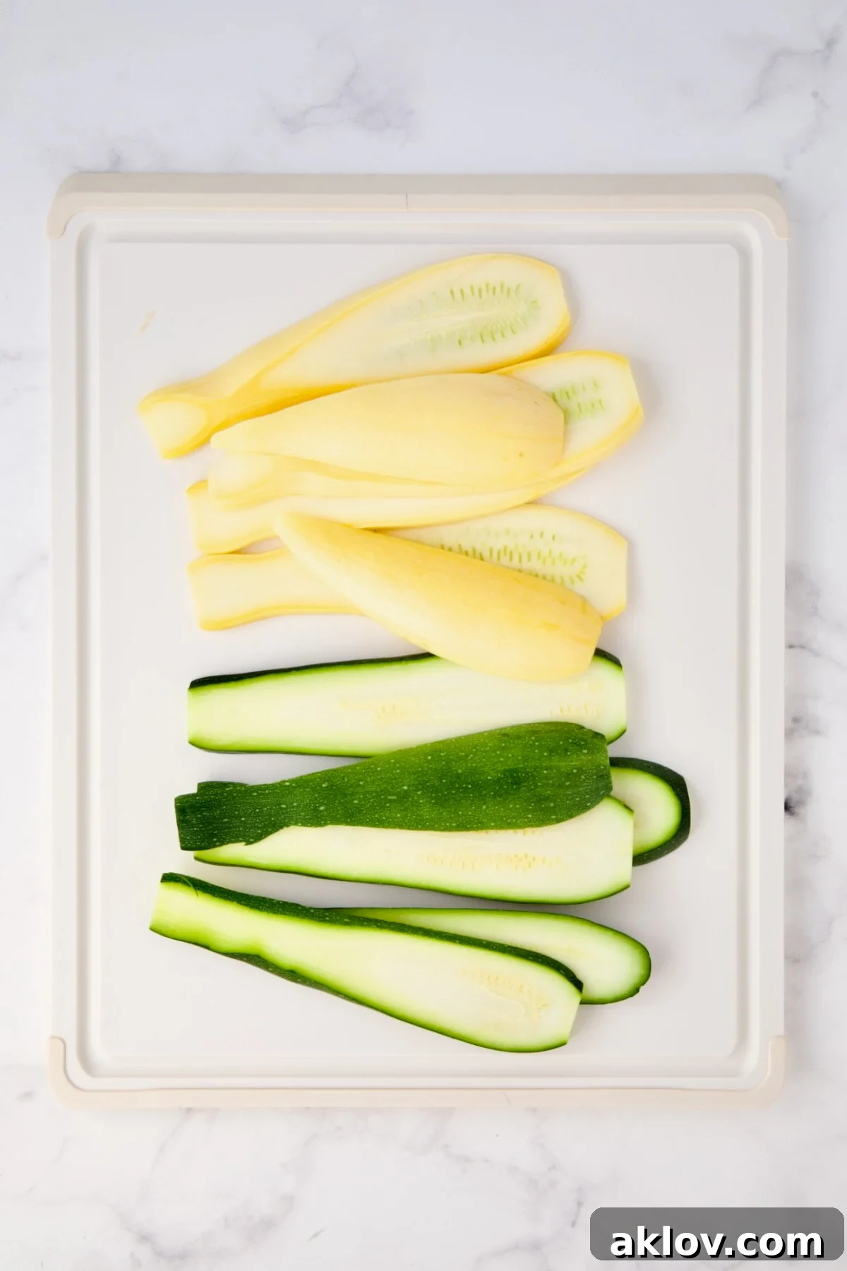 Sliced yellow squash and zucchini on white plastic cutting board.