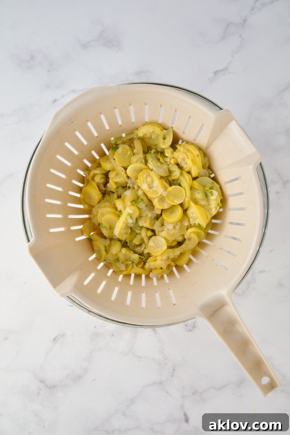 Cooked yellow squash, onion and jalapeno in beige colander set over a glass bowl.