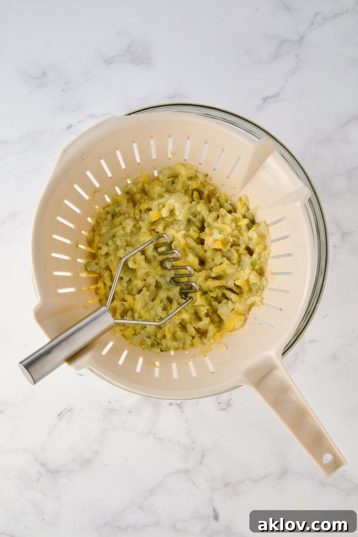 Cooked mashed yellow squash, onion and jalapeno in beige colander set over a glass bowl with potato masher.