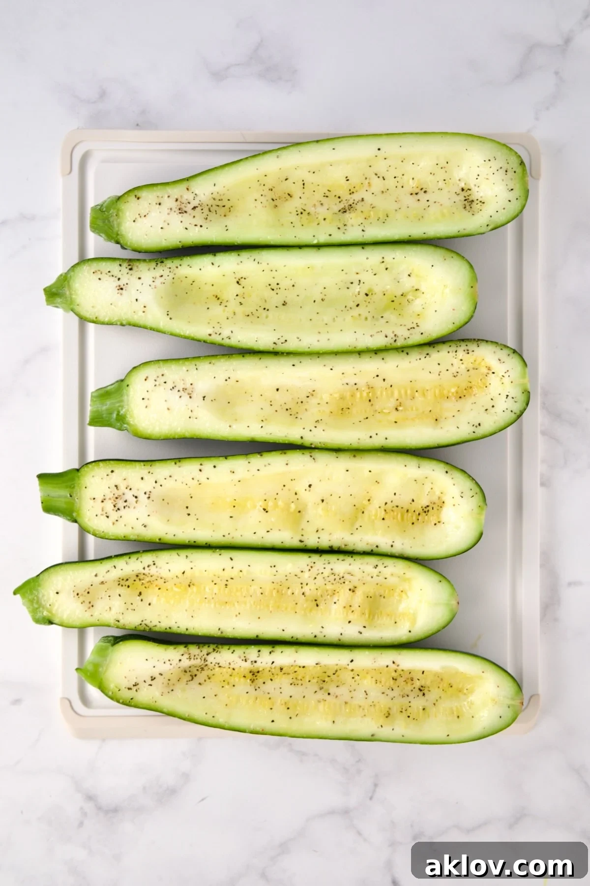 Six hollowed out zucchini halves on white plastic cutting board seasoned with salt and black pepper.