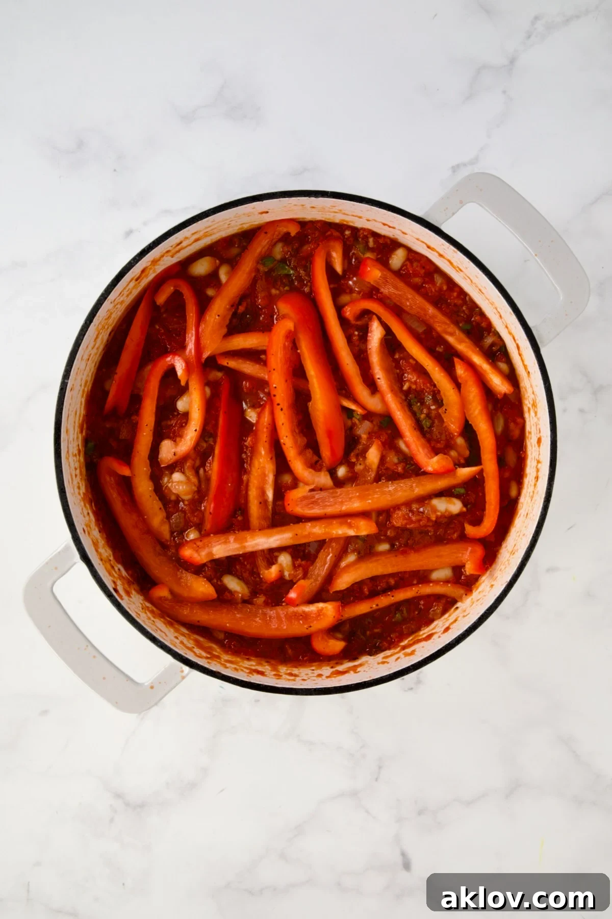 Red bell pepper slices placed over the top of tomato sauce with beans in white enamel cast iron skillet.