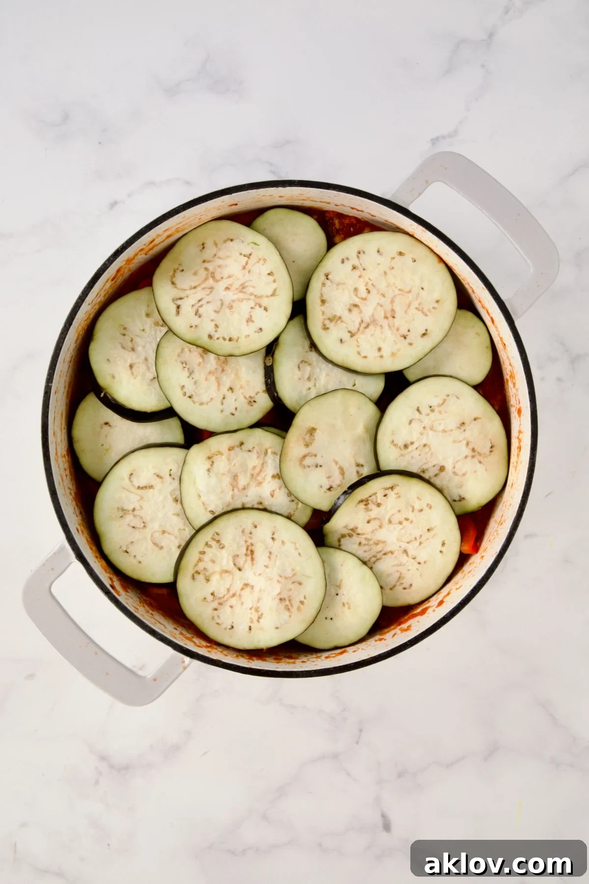 Eggplant slices placed over red bell pepper in white enamel cast iron skillet.