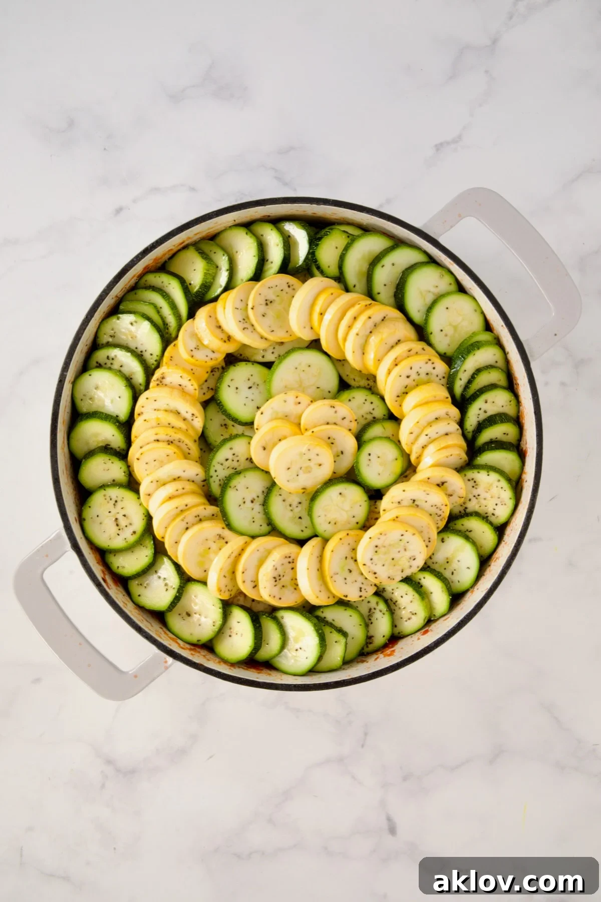 Zucchini and yellow squash slices placed over eggplant in a circular pattern in white enamel cast iron skillet.