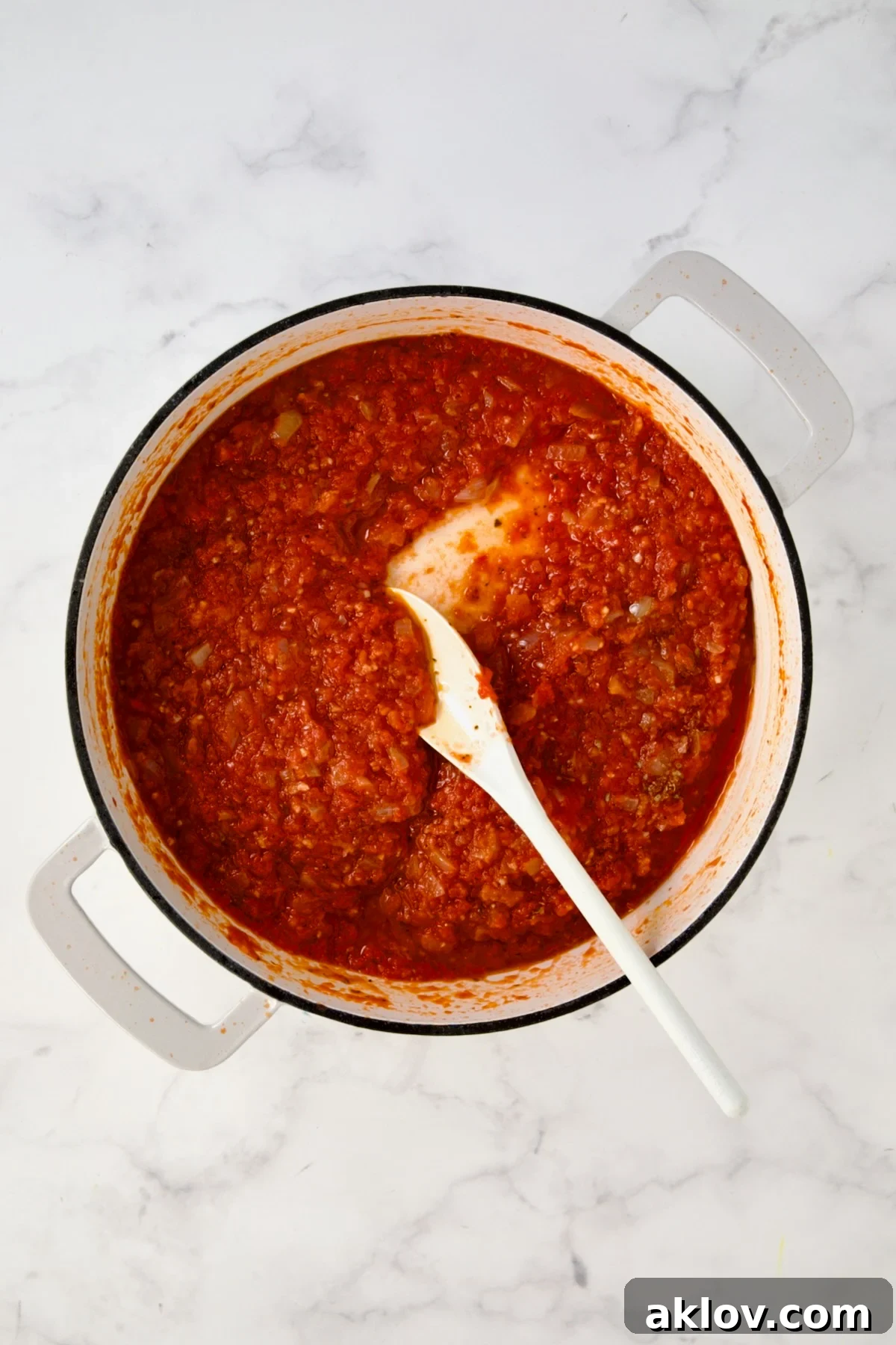 White plastic spoon stirring tomato sauce in white enamel cast iron skillet.